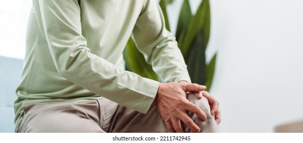 Asian Senior Man Feel Knee Pain Ache. Older Mature Male Hurt At Knee While Sitting On Sofa In Living Room At Home. Bone Problam Health Care And Medical Insurance Concept.