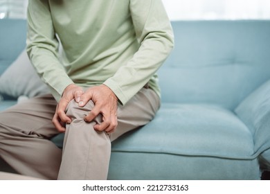 Asian Senior Man Feel Knee Pain Ache. Older Mature Male Hurt At Knee While Sitting On Sofa In Living Room At Home. Bone Problam Health Care And Medical Insurance Concept.