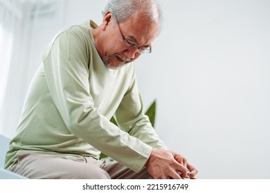 Asian Senior Man Feel Knee Pain Ache. Older Mature Male Hurt At Knee While Sitting On Sofa In Living Room At Home. Bone Problam Health Care And Medical Insurance Concept.