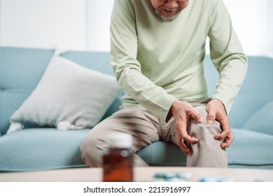 Asian Senior Man Feel Knee Pain Ache. Older Mature Male Hurt At Knee While Sitting On Sofa In Living Room At Home. Bone Problam Health Care And Medical Insurance Concept.