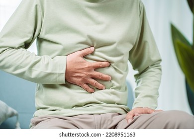 Asian Senior man feel stomachache. Older mature male touch belly while sitting on sofa in living room at home. Bowel problem health care and medical insurance concept. - Powered by Shutterstock