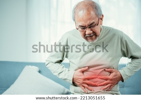 Asian Senior man feel stomachache. Older mature male touch belly while sitting on sofa in living room at home. Bowel problem health care and medical insurance concept.
