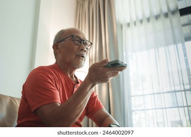 Asian Senior man in glasses sitting on a couch, enthusiastically giving voice commands on smartphone to his smart home system - Powered by Shutterstock