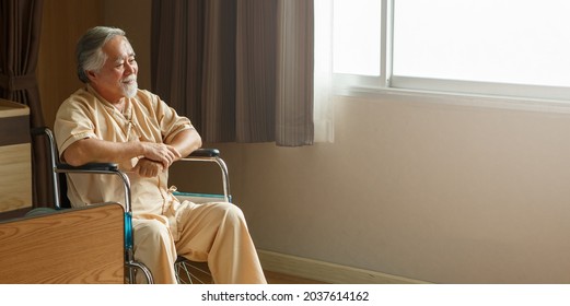 Asian Senior Man Patient Sit On Wheelchair  Alone And Looking Out The Window At Hospital Ward . Lonely Old Male  In Nursing Home. Loneliness Elder. Depressed Mature . Copy Space