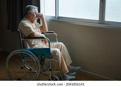 asian senior man patient sit on wheelchair holding head with hands alone and looking out the window at hospital ward . Lonely old male  in Nursing home. loneliness elder. Depressed mature . 