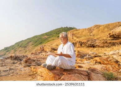 Asian senior man practicing meditation with ocean nature on rocky coastal hill at summer sunset. Retirement elderly people do outdoor relaxing yoga exercise. Mental health care and motivation concept. - Powered by Shutterstock