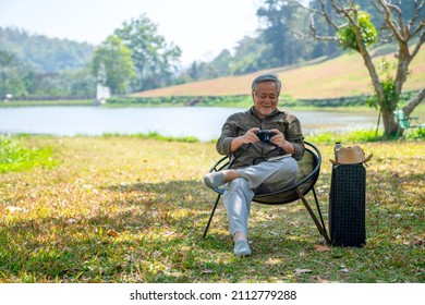 Asian Senior Man Sit On Outdoor Chair Using Digital Camera Photography Beautiful Nature At Public Park. Healthy Elderly Retired Male Relax And Enjoy Outdoor Leisure Activity Hobby In Summer Sunny Day.