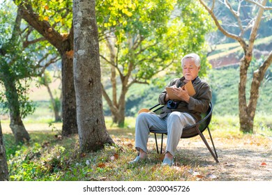 Asian Senior Man Sitting Alone On Outdoor Chair In Nature Park With Writing Or Drawing On Paper Notebook. Healthy Elderly Retired Male Relax And Enjoy Outdoor Leisure Activity Hobby In Sunny Day