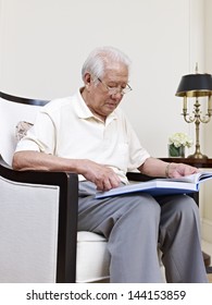 Asian Senior Man Sitting In An Armchair And Reading.