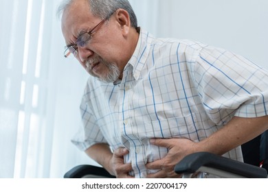 Asian Senior Man Sitting On Wheelchair Having Stomachache. Hand Holding Tummy To Relieve Pain. Senior Male Patient Suffering From Sickness