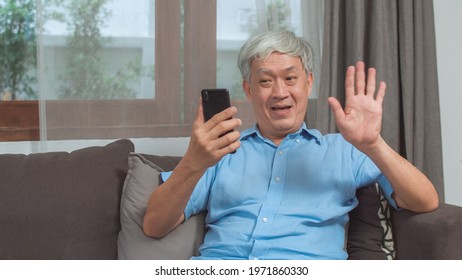 Asian Senior Man Video Call At Home. Asian Senior Older Chinese Male Using Mobile Phone Video Call Talking With Family Grandchild Kids While Lying On Sofa In Living Room At Home Concept.