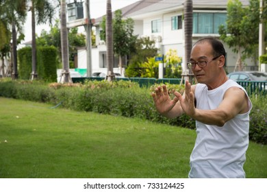 Asian Senior Man Wear White Shirt Stand And Practice Tai Chi On The Grass In The Garden