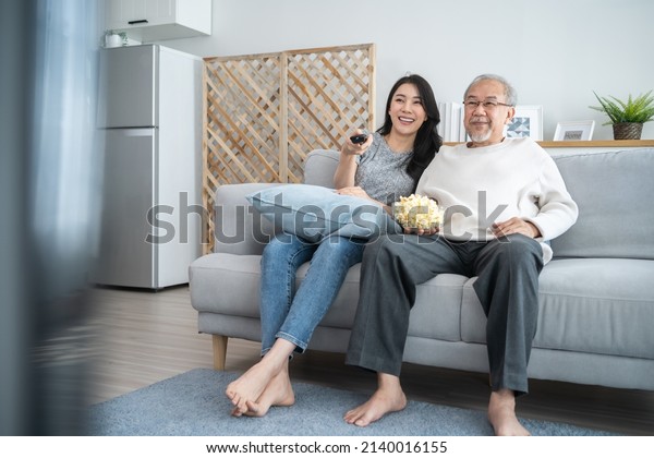 Asian senior mature man and young daughter sitting\
on sofa watch movie. Attractive beautiful girl spend time with\
elderly father enjoy comedy show on TV and eat popcorn together in\
living room at home
