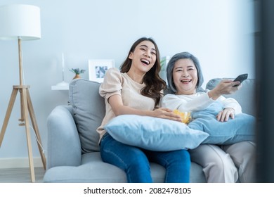 Asian Senior Mature Woman And Daughter Sit On Sofa Watch Funny Movie. Loving Elderly Older Mother Spend Time In Living Room At Home With Beautiful Girl Enjoy Comedy Show On TV And Eat Snacks Together.