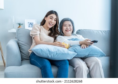 Asian Senior Mature Woman And Daughter Sit On Sofa Watch Funny Movie. Loving Elderly Older Mother Spend Time In Living Room At Home With Beautiful Girl Enjoy Comedy Show On TV And Eat Snacks Together.