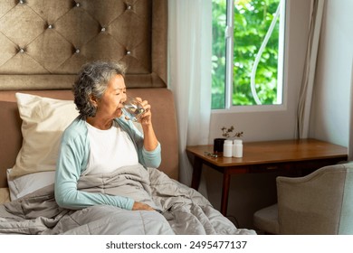 Asian senior mature woman drinking water from a glass after wake up on the bed in the morning. Elderly retired woman taking medicine or vitamins complex supplement at home. Healthcare concept. - Powered by Shutterstock