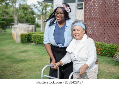 Asian Senior Mother Walk With Walker And African American Carer Support In Garden At Home. Old Woman Disabled Walking And Black Caregiver Young Woman Helping In Back Yard Park Outdoors. Health Care 