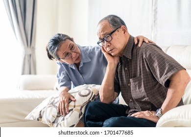 Asian senior old man having headache stress problem put hand on temple sitting on sofa at home. Elder wife sit beside consoling husband put hug, put hand on shoulder. Make comfort talking to relax him