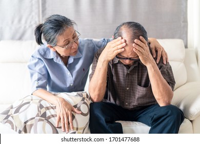 Asian senior old man having headache stress problem put hand on temple sitting on sofa at home. Elder wife sit beside consoling husband put hug, put hand on shoulder. Make comfort talking to relax him