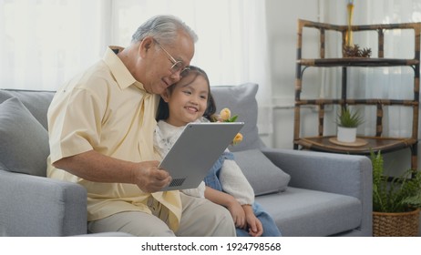 Asian Senior Old Man Looking To Tablet Computer And Granddaughter Come Visitor At Home, Grandfather Reading News On Digital Tablet With His Kid On Sofa In Living Room