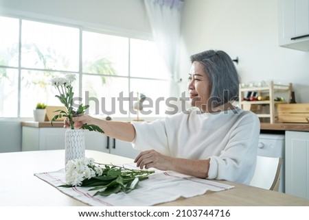 Asian senior old woman puts flowers on vase with happiness in kitchen. Beautiful active elderly grandmother sit on table, feeling happy while decorate and renovate apartment. Activity at home concept.