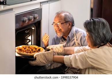 Asian Senior Older Couple Grandparents Making Pizza In Kitchen At Home. Aged Woman Open Oven And Bring Food Out From Machine. Elder Man Look At Meal And Smell With Smile Enjoy Famiy Activity Together.