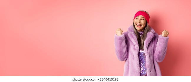 Asian senior woman celebrating, cheering and winning, making fist pump signs in triumph, achieve goal and smiling satisfied, standing in stylish faux fur coat, pink background. - Powered by Shutterstock