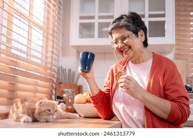 Asian senior woman drink a cup of coffee with domestic cat at home. Attractive elderly grandma feeling happy and relax, enjoy spending leisure time with brown furry Persian kitty animal in kitchen. - Powered by Shutterstock