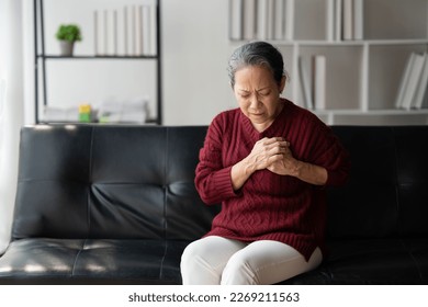 Asian senior woman feeling heart pain and holding her chest while sitting on the sofa in her living room.