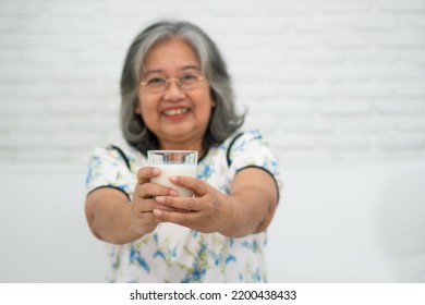 Asian Senior Woman Holding Glass Milk While Relaxing On A Sofa Living Room For Retirement Wellness. Elderly Woman Drinking A Glass Of Milk To Maintain Her Wellbeing. Concept Of Wellness And Healthy.