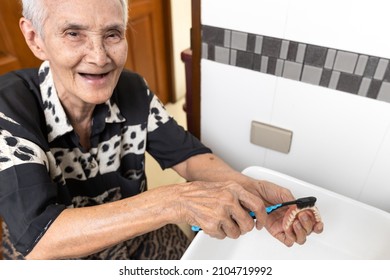 Asian senior woman holding a removable denture,old elderly brushing her denture plate with a toothbrush at the sink in bathroom,clean her false teeth for oral hygiene,tooth care,dental health concept
