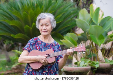 Asian senior woman holding ukulele while standing in a garden. Relaxing by singing and play small guitar happy and enjoy life after retired. Concept of old people and healthcare.