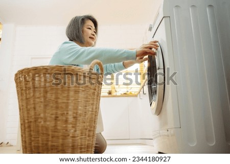 Asian Senior woman housekeeper loading clothes to washing machine. Routine activity in house concept.