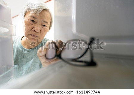 Asian senior woman with memory impairment symptoms,forget her glasses in the refrigerator or storing glasses in the fridge,female elderly having dementia, cognitive impairment, alzheimer's, amnesia