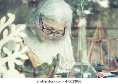 Asian Senior Woman Painting At Her Home Studio, View Through Window. Soft Focus On Eyes. Active Old Lady Concept