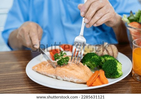 Asian senior woman patient eating Salmon steak breakfast with vegetable healthy food while sitting and hungry on bed in hospital.