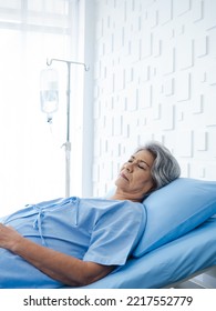 Asian Senior Woman Patient Grey Hair Sleeping On Bed In Modern White Hospital Room, Vertical Style. An Old Lady Is Sick And Lying On The Patient Bed, Recovering On Saline Drip Solution.