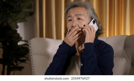 Asian senior woman sitting on sofa, talking on her phone with a worried and concerned expression, deeply engaged in her conversation. - Powered by Shutterstock