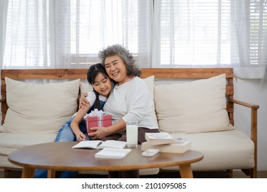 Asian Senior Woman Snuggle Little Girl With Love And Smile On Couch In A Living Room After Receiving Red Present Box. Granddaughter Visiting Grandmother At Home. Family Insurance Concept. Copy Space