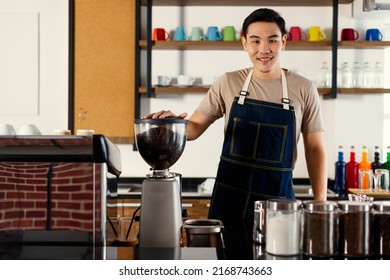 Asian short black hair young successful handsome male barista wear brown shirt and blue jeans bib apron stand smiling behind counter bar full of paper coffee cups, maker machine, beans and equipments.