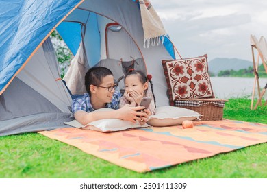 Asian siblings enjoying camping by lake, sharing smartphone in colorful tent. Boy and girl laughing together on picnic blanket. Family bonding, outdoor adventure, and technology in nature concept. - Powered by Shutterstock