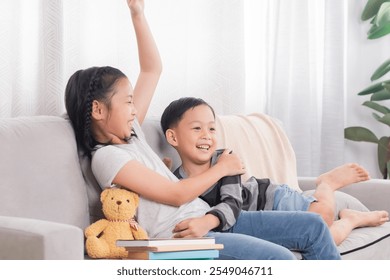 Asian siblings having fun together on sofa. Younger boy playfully lying on girl, both laughing and smiling. Casual and joyful moment, showcasing strong sibling relationship in cozy home setting. - Powered by Shutterstock