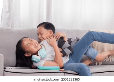 Asian siblings having fun together on sofa. Younger boy playfully lying on girl, both laughing and smiling. Casual and joyful moment, showcasing strong sibling relationship in cozy home setting. - Powered by Shutterstock