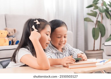 Asian siblings laughing together while using digital tablet. Young boy and girl sitting at table watch cartoons at home. Bright indoor setting, joyful expression, sibling bonding, technology use. - Powered by Shutterstock