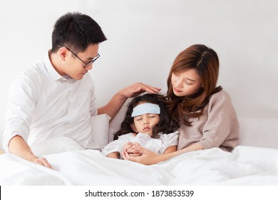 Asian Sick Girl Sleeping On The Bed With Fever Gel Pack On Her Forehead. Father And Mother Take Care Of Their Little Young Daughter Who Got A Fever From Flu Virus. Sick Little Kid Laying On The Bed.