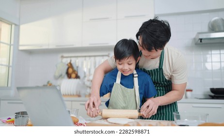 Asian single father teaching little son making dough for bread, learning from videos on social media using labtop in the kitchen - Powered by Shutterstock