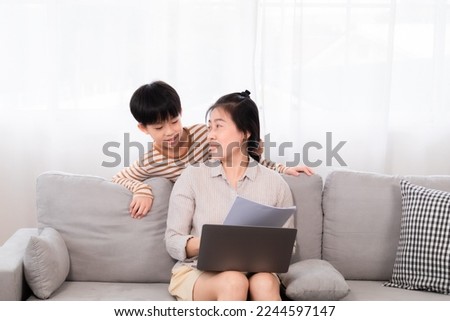 Asian single mom sitting on sofa disturbed by her son, tease mother's  while she focused on typing and working remotely at home on laptop. Children demand attention from the family.