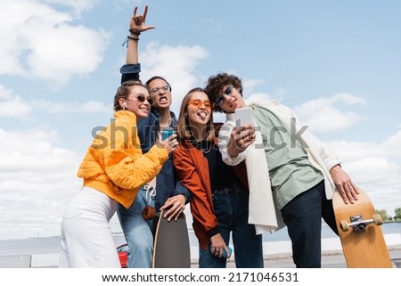 asian skater showing rock sign near excited friends taking selfie outdoors