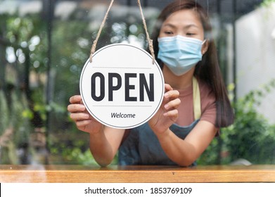 Asiatischer Kleinunternehmer, der eine Schutzmaske trägt und ein Drehzeichen trägt, um sich nach der Quarantäne aufgrund einer Koronavirus-Pandemie wieder zu öffnen. Frau hängt offenes Schild auf dem Glasfenster. Konzentriere dich auf Schild.