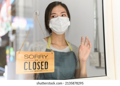 Asian small business owner wearing protective face mask and turning sign to close after the quarantine due to coronavirus pandemic. Woman hanging close sign on the glass window. Focus on sign.